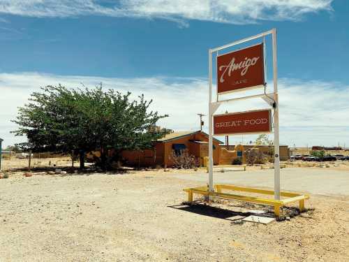 A weathered sign for Amigo Café stands in a dusty area, with a tree and a small building in the background.