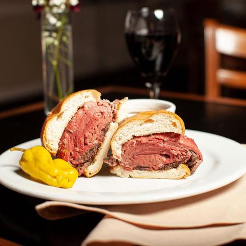 A sliced roast beef sandwich on a plate, accompanied by a pepper and a small bowl of sauce, with a glass of red wine in the background.