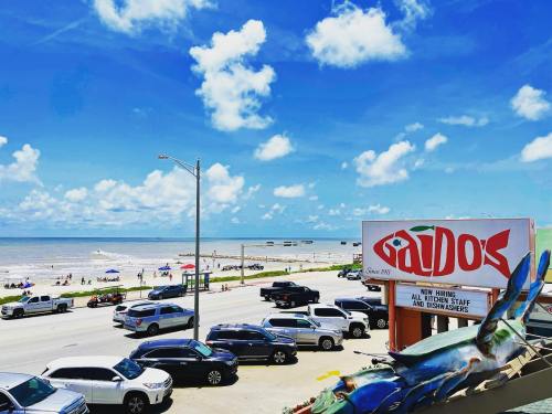 A beach view with cars parked along the road, a restaurant sign, and people enjoying the sandy shore under a blue sky.