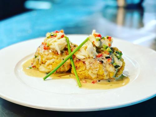 Two crab cakes topped with fresh crab meat and garnished with green onions and red peppers, served on a white plate.