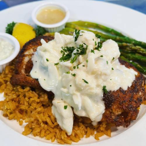 A plate of seasoned fish topped with creamy crab sauce, served with rice, asparagus, and lemon wedges.