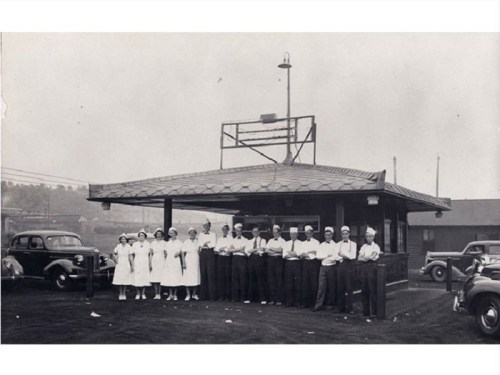A historic black-and-white photo of a group of people in uniforms outside a small building, with vintage cars nearby.