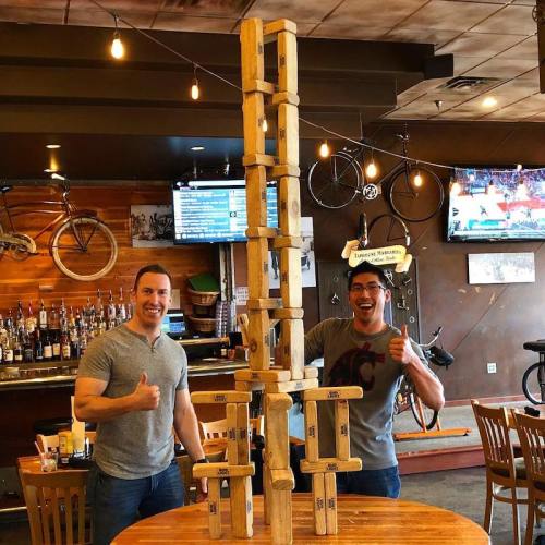 Two men give thumbs up next to a tall wooden block tower in a restaurant with bikes and TVs in the background.