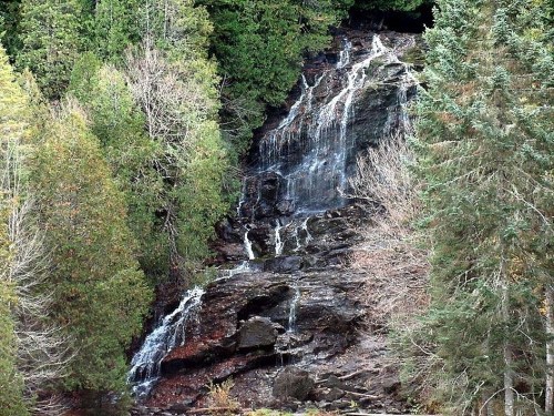 A cascading waterfall flows down rocky terrain, surrounded by lush green trees and autumn foliage.