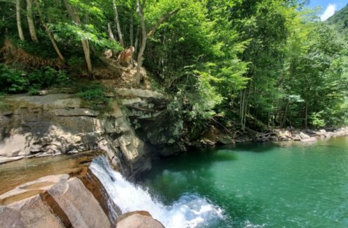 A serene waterfall cascades into a clear green pool, surrounded by lush trees and rocky terrain.