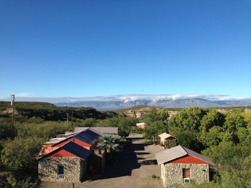 A scenic view of stone buildings with red roofs, surrounded by greenery and mountains under a clear blue sky.