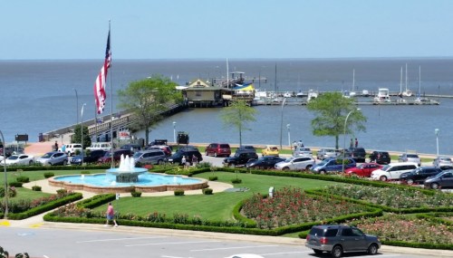 A waterfront view featuring a fountain, landscaped gardens, parked cars, and a pier with boats in the background.