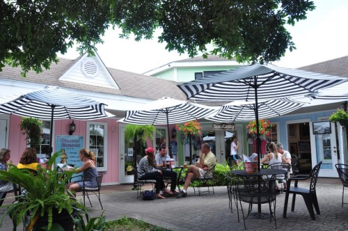 A vibrant outdoor café scene with people sitting under striped umbrellas, surrounded by colorful buildings and greenery.