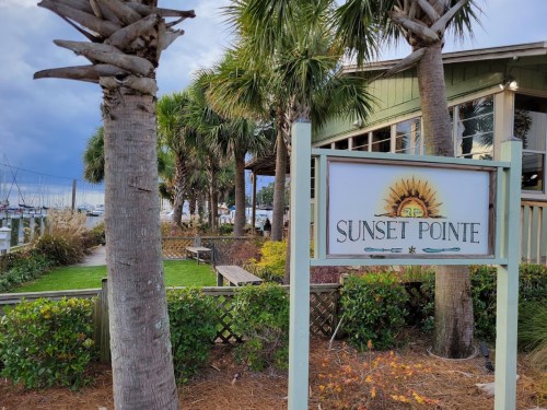 Sign for Sunset Pointe with palm trees and a marina in the background, showcasing a scenic waterfront location.