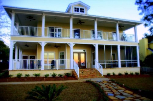 A large yellow house with multiple balconies, white railings, and a stone pathway in front, surrounded by greenery.