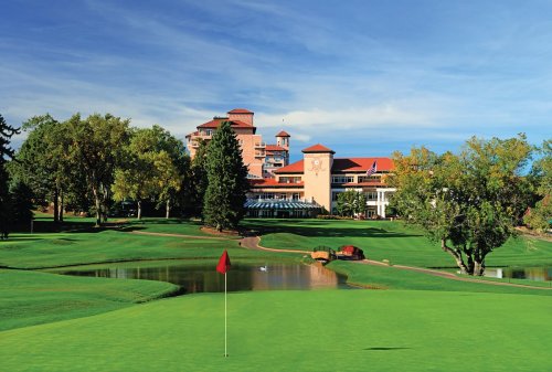 A scenic golf course with a red flag, lush green grass, a pond, and a resort building in the background under a blue sky.