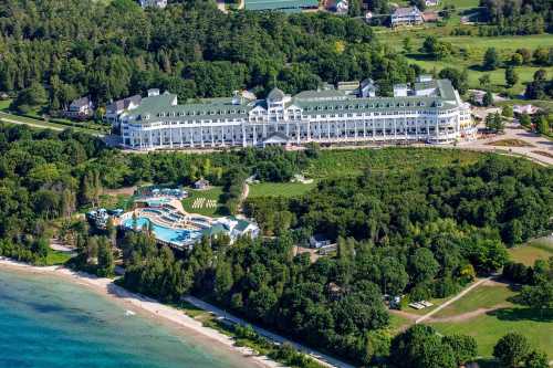 Aerial view of a large hotel surrounded by greenery and a pool area near a sandy beach and blue water.