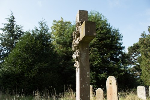 A weathered stone cross in a cemetery, surrounded by tall trees and gravestones under a clear blue sky.