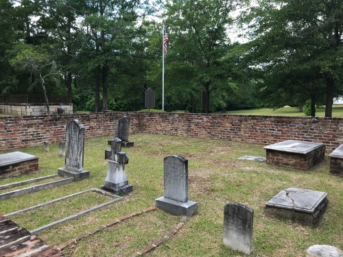 A small, walled cemetery with several gravestones and a flagpole in the background, surrounded by trees.