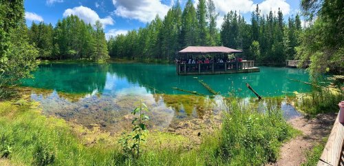 A serene lake surrounded by lush greenery, with a wooden platform and people enjoying the view under a blue sky.