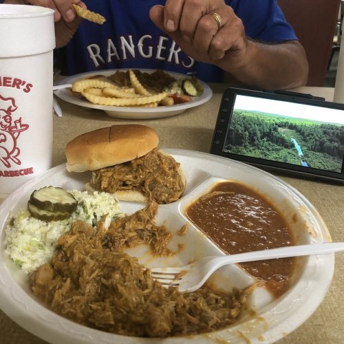 A plate of pulled pork with coleslaw, pickles, and beans, alongside a drink and a phone displaying a forest view.