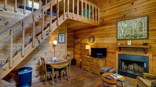 Cozy wooden cabin interior with a staircase, dining table, TV, and fireplace, featuring warm tones and rustic decor.