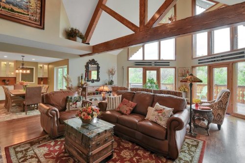 Cozy living room with leather sofas, wooden beams, large windows, and a rustic coffee table adorned with flowers.