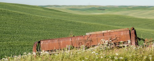 Rusty machinery rests in a grassy field, surrounded by rolling green hills under a clear sky.
