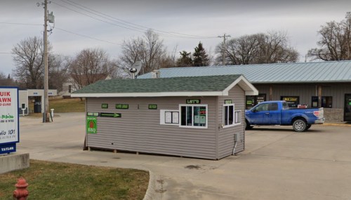 A small BBQ stand with a sign, next to a building and a blue pickup truck parked nearby. Trees in the background.