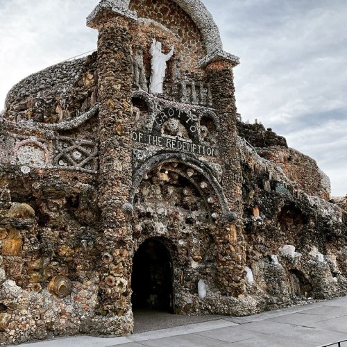 A stone structure adorned with shells and sculptures, featuring a statue of a figure at the top, under a cloudy sky.