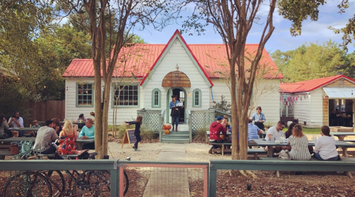 A charming café with a red roof, surrounded by outdoor seating and trees, bustling with people enjoying their time.