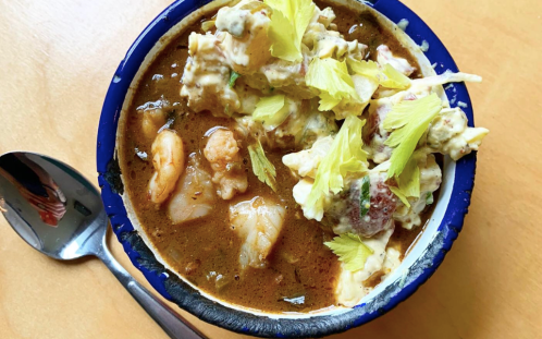 A bowl of shrimp stew topped with celery, served with a spoon on a wooden surface.