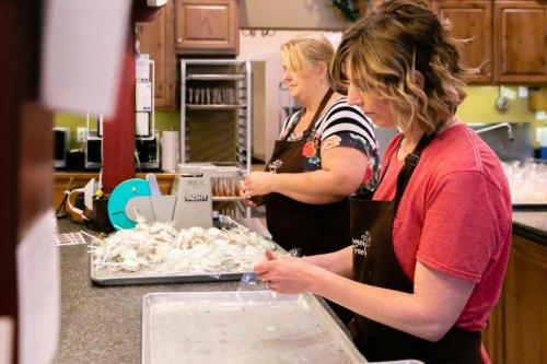 Two women in aprons prepare packaged items at a kitchen counter, with bags and a sealing machine in the background.