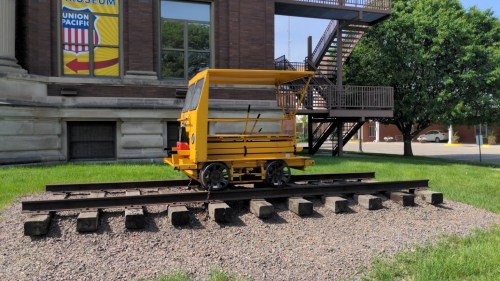 A yellow rail cart sits on tracks outside a museum, with a staircase and greenery in the background.