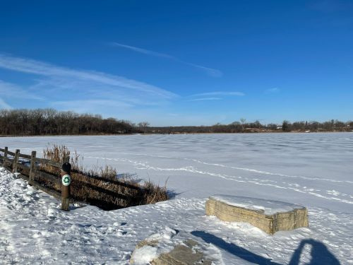 A snowy landscape by a frozen lake, with a wooden fence and clear blue sky in the background.
