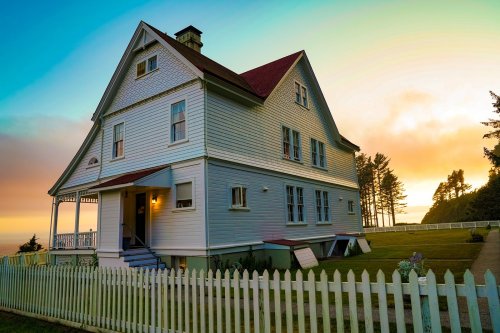 A charming two-story house with a red roof, surrounded by a white picket fence, at sunset with trees in the background.