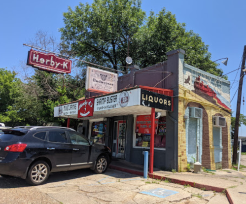 A small storefront with signs for "Harby-K" and "Liquors," featuring a parked car and trees in the background.