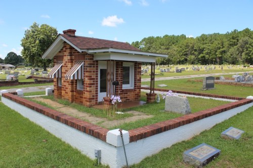 A small brick house-shaped tomb in a cemetery, surrounded by grass and gravestones under a clear blue sky.