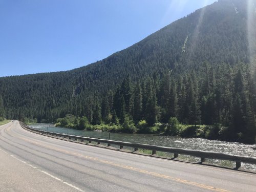 A winding road alongside a river, surrounded by lush green trees and a mountain under a clear blue sky.