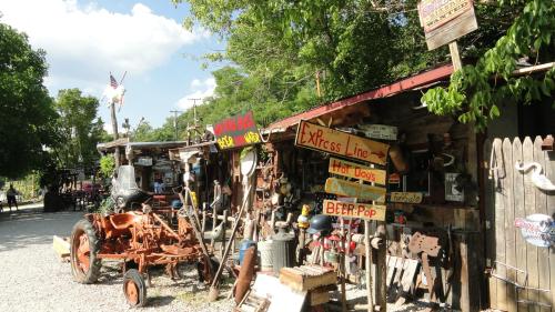A rustic roadside stand with colorful signs, an old tractor, and various vintage items surrounded by trees.