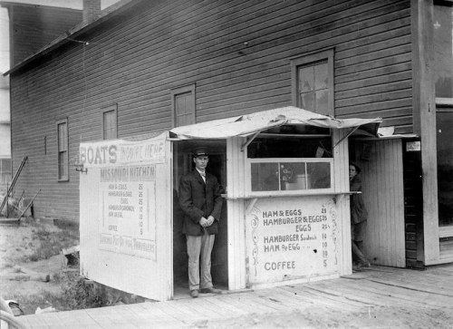 A historical black-and-white photo of a man standing outside a small food stand with a menu board.
