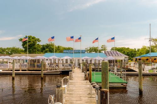 A wooden dock leads to a seafood restaurant with American flags and a blue roof, surrounded by calm water and greenery.