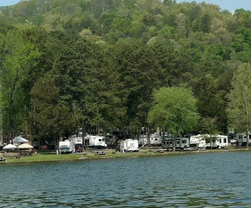A serene lakeside view with RVs parked among tall trees and green hills in the background.