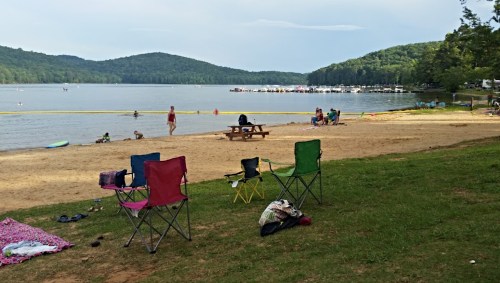 A sandy beach by a lake with colorful chairs, people swimming, and boats in the distance surrounded by green hills.