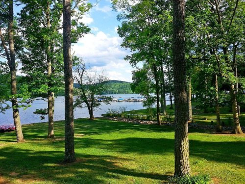 A serene lakeside view with lush green trees, a calm water surface, and distant hills under a partly cloudy sky.