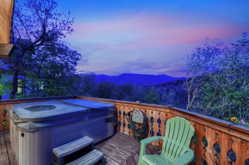 A serene view from a deck featuring a hot tub and a green chair, with mountains and a colorful sunset in the background.