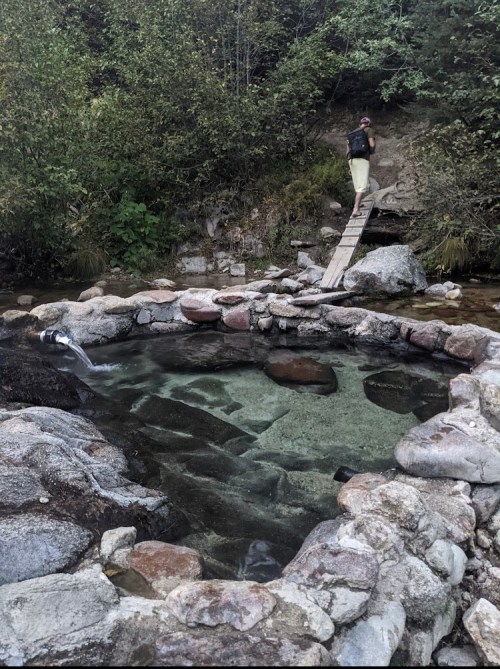 A natural hot spring surrounded by rocks, with a wooden bridge leading to a person standing nearby in a lush setting.