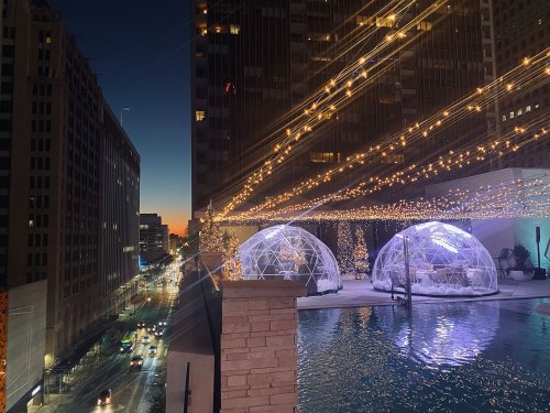 A rooftop view at dusk featuring illuminated igloos, string lights, and a city skyline in the background.