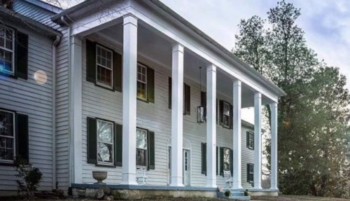A large white house with tall columns, green shutters, and a porch surrounded by trees.