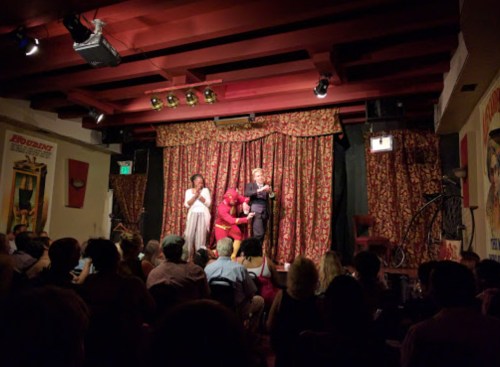 A lively performance on stage with two performers and an audience seated in a cozy, decorated venue.