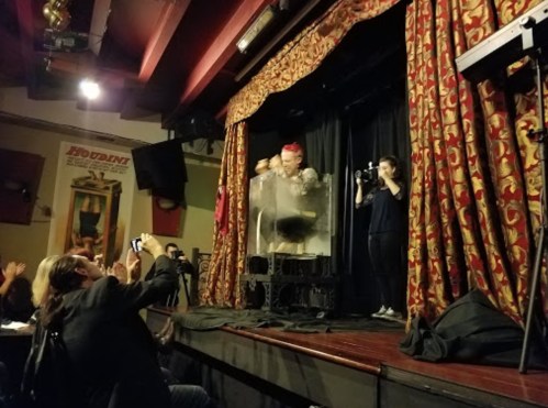 A performer in a transparent box on stage, with an audience member taking photos and another person playing a trumpet.