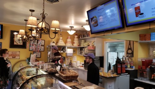 A cozy café interior with a display case, menu screens, and staff serving customers. Warm lighting and decor create a welcoming atmosphere.