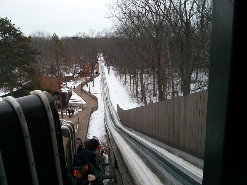 View from the top of a snowy toboggan run, with people waiting and a winding track visible in the distance.