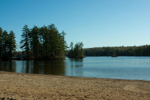 A serene lake scene with sandy shore, surrounded by trees and small islands under a clear blue sky.
