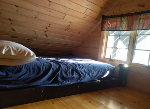 Cozy attic bedroom with wooden walls, a bed covered in a blue blanket, and sunlight streaming through the windows.